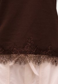 Καφέ ύφασμα με λουλουδένια δαντέλα πάνω από άσπρο παντελόνι με ορατό κορδόνι, που δείχνει υφή και λεπτομέρειες στρώσεων.