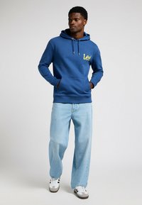 Sudadera azul con capucha y logo amarillo "Lee", bolsillo tipo canguro en el frente, puños acanalados, combinada con jeans de pierna ancha de un azul claro y zapatillas blancas.