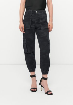 Femme portant un pantalon cargo jogger camouflage noir avec des poignets élastiques et des sandales noires à talons hauts avec bride à la cheville, debout sur un fond blanc.