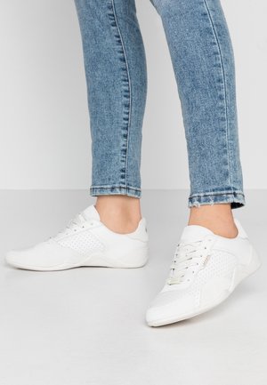 Person wearing light blue jeans and white Lacoste low-top sneakers standing on a light gray floor against a white wall.