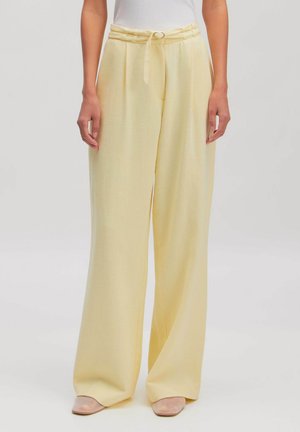 Vrouw draagt lichtgele, wijde broek met hoge taille en bijpassende riem en nudekleurige schoenen tegen een effen lichte achtergrond.