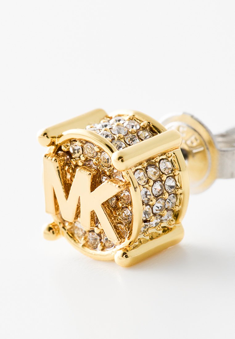 Boucle d'oreille clou en tone doré, de forme ronde, incrustée de cristaux transparents, et portant au centre un logo "MK" en évidence.