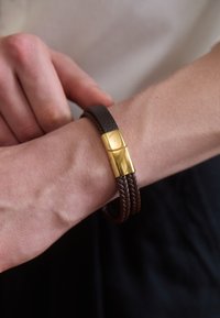 Brățară din piele împletită maro cu un închizător de culoare aurie, având o suprafață netedă și un detaliu de design rectangular pentru o închidere sigură.