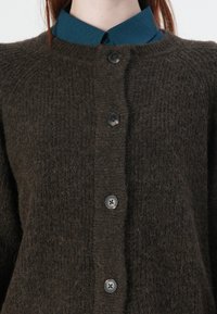 Dark brown knitted cardigan featuring a textured finish, five black buttons, and a collar from a blue shirt underneath.