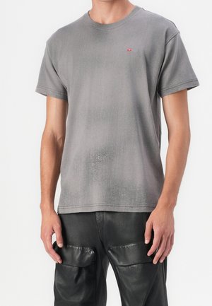 Homme portant un t-shirt gris à manches courtes avec un petit logo rouge sur la poitrine et un pantalon en cuir noir avec de grandes poches avant.