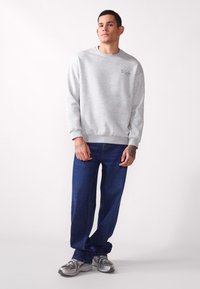 Grå hettegenser med ribbestrikkede mansjetter og kant, med tekst på venstre bryst; kombinert med blå denimjeans og sølvfargede treningssko.