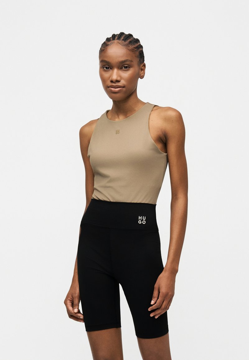 Γυναίκα με πλεξούδες, φορώντας μπεζ αμάνικο τοπ και μαύρο biker σορτς ψηλής μέσης με λογότυπο "HUGO" στη μέση.