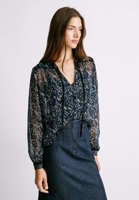 Blusa trasparente blu marino con un motivo floreale in tonalità di blu e bianco, dotata di chiusura con bottoni e cappuccio regolabile, abbinata a una gonna di denim.