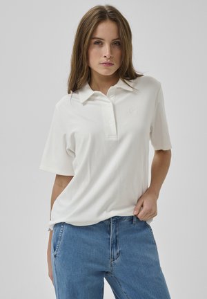 Mladá žena nosiaca biele polo tričko s krátkym rukávom a modré džínsy, stojaca s jednou rukou v vrecku na jednoliatom pozadí.