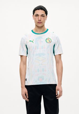 Jonge man draagt een wit Senegal voetbalshirt met een groene kraag en het Puma-logo, staand voor een effen witte achtergrond.