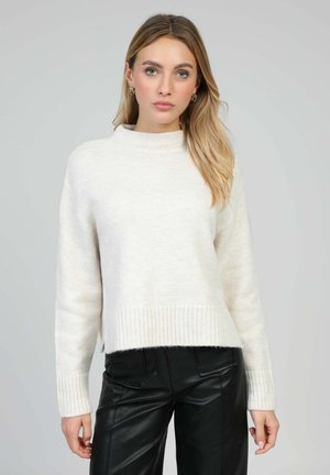 Femme aux longs cheveux blonds portant un pull en maille blanc et un pantalon en cuir noir, debout devant un fond gris clair uni.