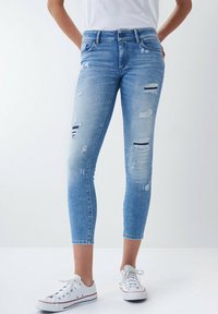 Jeans ajustados de mezclilla azul claro con detalles desgastados y parches deshilachados. Presentan una cintura de altura media y un corte a la altura del tobillo. Combinados con zapatillas blancas.
