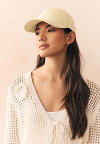 Yellow baseball cap made of fabric, featuring a curved brim and stitched seams. Model wears a white knit cardigan with a floral accent.