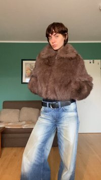 Brązowa krótka kurtka z futerału sztucznego z wysokim kołnierzem, połączona z jasnoniebieskimi szerokimi spodniami dżinsowymi z wyblakłym wzorem.