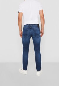 Vaqueros de mezclilla azul de corte ajustado con una textura desgastada, cinco bolsillos y sutilezas en las costuras en la parte trasera. Combinados con una camiseta blanca corta.