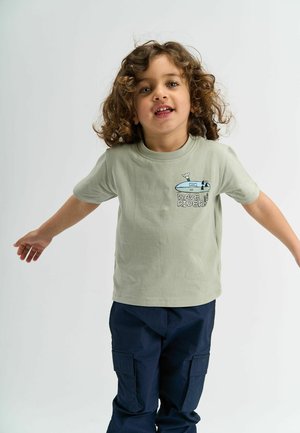 Svetlozelené bavlnené tričko s potlačou surfového dosky a textom "WAVE RIDER." Skombinované s tmavomodrými nohavicami cargo s bočnými vreckami.