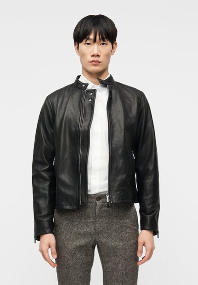 Jeune homme portant une veste en cuir noire sur une chemise blanche boutonnée et un pantalon gris, debout devant un fond blanc uni.