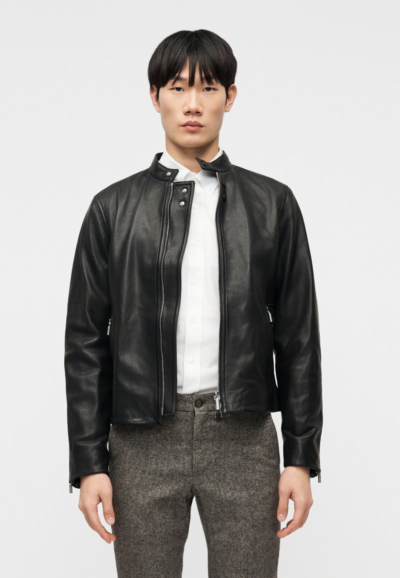 Νεαρός άνδρας φοράει μαύρο δερμάτινο μπουφάν πάνω από λευκό πουκάμισο με κουμπιά και γκρι παντελόνι, στέκεται μπροστά σε ένα απλό λευκό φόντο.