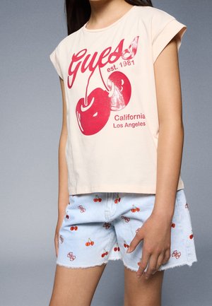 Kind trägt beiges ärmelloses Top mit rotem Kirschmotiv und dem Text „Guess est. 1981 California Los Angeles“ sowie hellblaue Shorts mit Kirschmotiven.