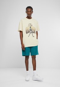 Cremefarvet grafisk t-shirt med "The Greatest" print, parret med teal shorts, hvide sneakers og synlige hvide sokker. Enkel, afslappet design.