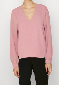 Rosa långärmad blus med V-ringning, tillverkad av textilerad tyg. Har samlade manschetter och en avslappnad passform, bärs med svarta byxor.