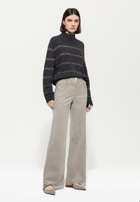 Frau steht in einem dunkelgrau gestreiften Pullover, beigen weit geschnittenen Hosen und taupefarbenenspitz zulaufenden Schuhen vor einem weißen Hintergrund.