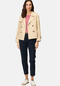 Beige dubbelknäppt trenchcoat med stora knappar, bärs över en röd och vit randig topp, ihop med marinblå byxor och vita sneakers.