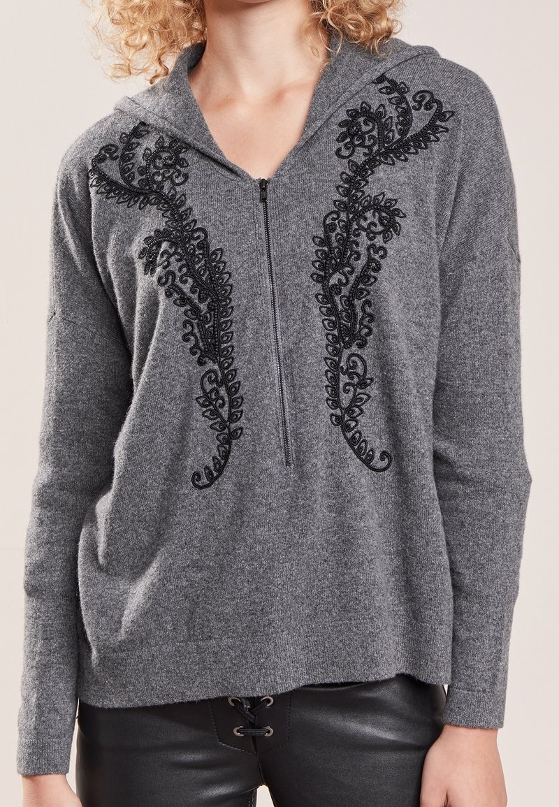 Sudadera gris hecha de una tela suave, con motivos florales bordados en negro a lo largo del frente, con cierre de cremallera y un ajuste relajado.