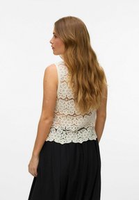 Krajkový top bez rukávů v barvě off-white s propracovanými květinovými vzory, s designem bez rukávů a vlnkovaným lemem, doplněný černou splývavou sukní.