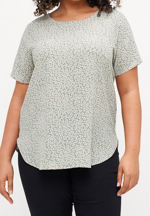 Vrouw draagt een grijs-groen shirt met korte mouwen en wit kiezelpatroon, gecombineerd met een zwarte broek, staand tegen een witte achtergrond.