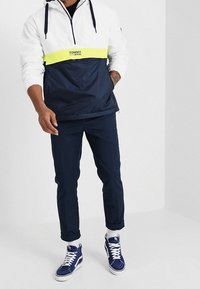 Pulloverjacka i vitt och marinblått, med ett gult rand och texten "TOMMY JEANS". Matchas med marinblå byxor och blå sneakers.