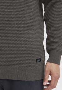 Maglione a maglia grigio scuro con un motivo a nido d'ape testurizzato, polsini a coste e una piccola etichetta sul lato inferiore con del testo.