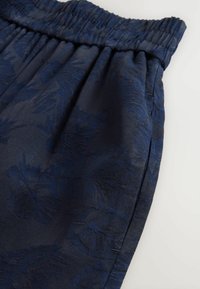 Marineblaue Shorts mit einem elastischen Bund, die ein strukturiertes florales Muster und eine glatte Oberfläche aufweisen, schräg fotografiert, um Fall und Designdetails zu zeigen.