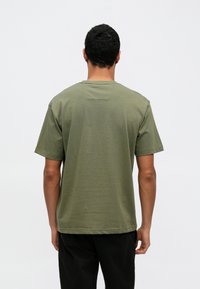 Oliiviroheline puuvillane T-särk lühikeste varrukatega, lõdva lõikega ja väikese silmuse detailiga kaela tagaosas. Sirge äärejoon.