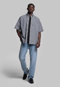 Camicia a maniche corte a righe in bianco e nero sopra una maglietta nera, abbinata a jeans azzurri e scarpe nere. Vestibilità casual e texture liscia.