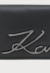 Mustrusega must nahast rahakott, millel on silmapaistev hõbedane logo kursiivis. Kompaktne disain, minimaalne õmbluste detail.