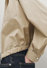 Light beige, cropped jacket with a smooth texture, elasticated hem, and buttoned cuffs, paired with dark denim pants featuring visible pockets.
