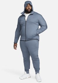 Light blue athletic tracksuit with a zip-up jacket featuring a hood, fitted pants, and white sneakers. Smooth fabric and minimal branding.