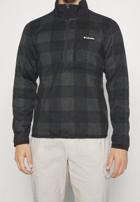 Homme portant une veste polaire Columbia noire et grise à carreaux avec une poche zippée sur la poitrine et un pantalon cargo beige, devant un fond uni.