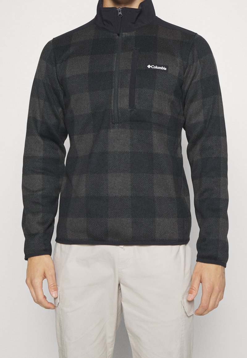 Homme portant une veste polaire Columbia noire et grise à carreaux avec une poche zippée sur la poitrine et un pantalon cargo beige, devant un fond uni.