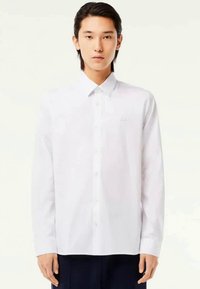 Camisa blanca de manga larga con cuello puntiagudo, cierre de botones y un logo en el pecho. Textura suave y corte estándar.