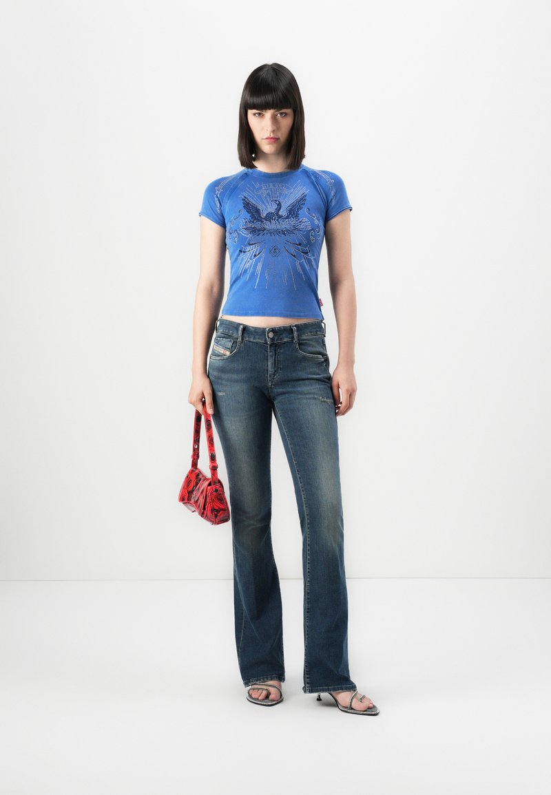 Woman with straight dark hair wearing blue graphic t-shirt, flare jeans, silver heeled sandals, holding small red patterned handbag against white background.