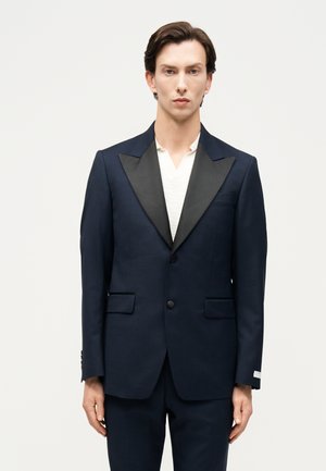 Mężczyzna ubrany w granatową marynarkę z czarnymi satynowymi klapami na białej koszuli, stojący na jednolitym jasnym tle.