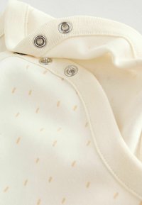 Krém színű baba body apró bézs pöttyös mintával. Elején patentos záródással és puha, rugalmas anyagból készült.