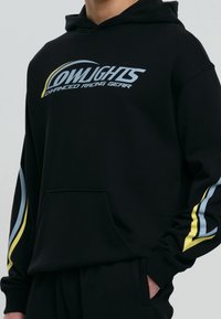 Czarna bluza z kapturem z przednią kieszenią, z niebieskim i żółtym napisem graficznym "LOWLIGHTS Enhanced Racing Gear" oraz kolorowymi akcentami na rękawach.