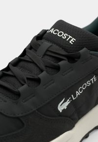 Black sneaker with textured upper, featuring a mesh layer, lace-up design, and white Lacoste logo with a crocodile accent. Lightweight sole.
