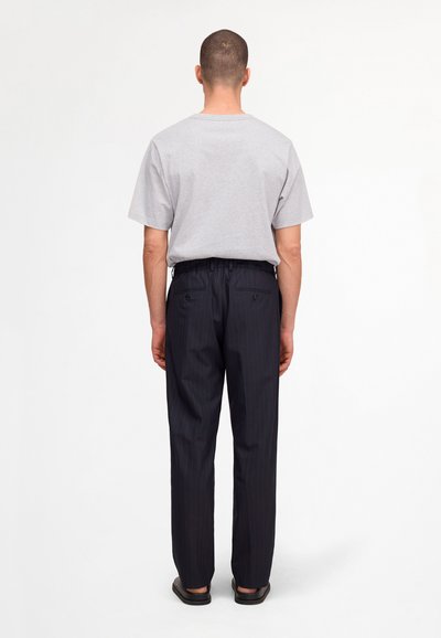 Homme aux cheveux courts portant un T-shirt gris clair, un pantalon sombre à fines rayures et des chaussures noires, debout de dos sur un fond blanc uni.
