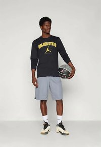 Fekete, hosszú ujjú póló "GOLDEN STATE" felirattal sárgában, szürke rövidnadrág és fekete sportcipő. Kosárlabdát tart, logós díszítéssel.