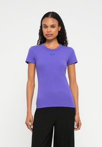 Donna dai capelli ricci che indossa una maglietta aderente a maniche corte di colore viola con un piccolo logo "HUGO", abbinata a pantaloni neri, in piedi su uno sfondo bianco.