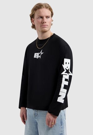 Blonde man met kort haar, gekleed in een zwart shirt met lange mouwen met een wit sterrenlogo op de borst en dikke witte tekst op de mouw, lichtblauwe spijkerbroek.